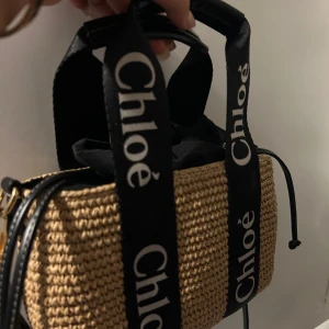 Beige väska från Chloé - Säljer en superfin beige väska från Chloé. Den är gjord i ett flätat material som ger en snygg och somrig look. Väskan har svarta handtag med Chloé-loggan tryckt i vitt, vilket ger en cool kontrast. Perfekt för både vardag och fest!