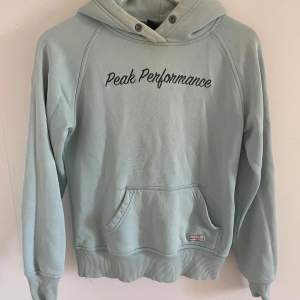 Säljer en snygg ljusblå hoodie från Peak Performance. Den har en klassisk design med en stor ficka fram och texten 'Peak Performance' broderad på bröstet. Huvan har metallöljetter för snörena och det finns en liten 'P' broderad på baksidan av huvan. Perfekt för en avslappnad stil!