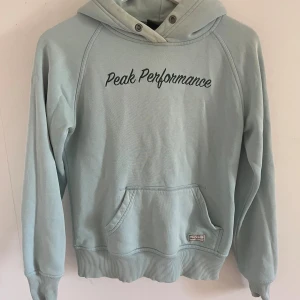 Ljusblå hoodie från Peak Performance - Säljer en snygg ljusblå hoodie från Peak Performance. Den har en klassisk design med en stor ficka fram och texten 'Peak Performance' broderad på bröstet. Huvan har metallöljetter för snörena och det finns en liten 'P' broderad på baksidan av huvan. Perfekt för en avslappnad stil!