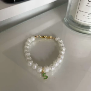 Pärlarmband med grön bokstavsberlock - Snyggt pärlarmband med vita pärlor och en grön bokstavsberlock 'S'. Armbandet har en guldfärgad kedja och lås. Perfekt för att ge en personlig touch till din outfit!