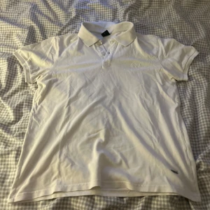 Vit pikétröja från Hugo Boss - Säljer en klassisk vit pikétröja från Hugo Boss. Den har en krage med knappar och är i ett mjukt bomullsmaterial. Perfekt för en stilren och avslappnad look.