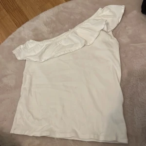 Vit oneshoulder topp från WE - Säljer en superfin vit oneshoulder topp från WE. Den har en snygg volangdetalj längs axeln och är perfekt för sommaren. Toppen är i storlek 158/164 och är i bra skick. Perfekt för en somrig dag eller en festkväll! 