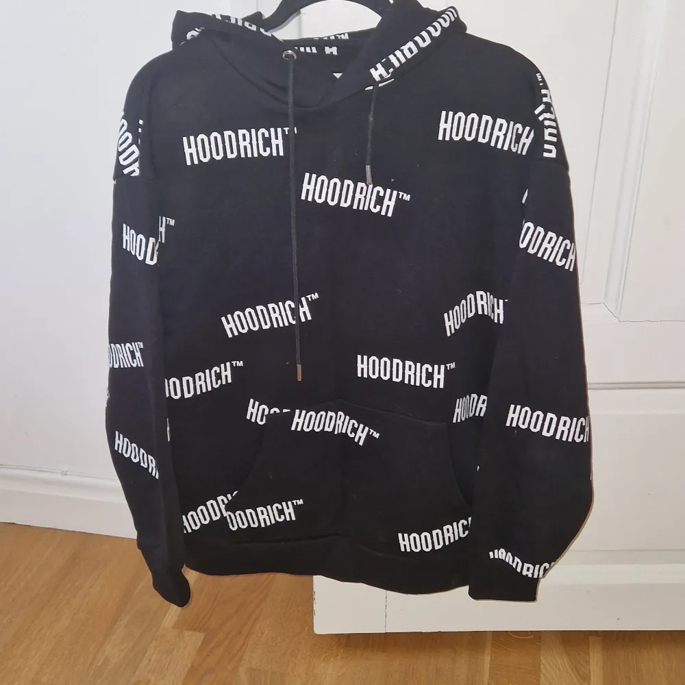 Säljer en svart hoodie från Hoodrich med vit text över hela tröjan. Den har en klassisk passform med en stor ficka fram och justerbar huva. Perfekt för en avslappnad stil!. Tröjor & Koftor.