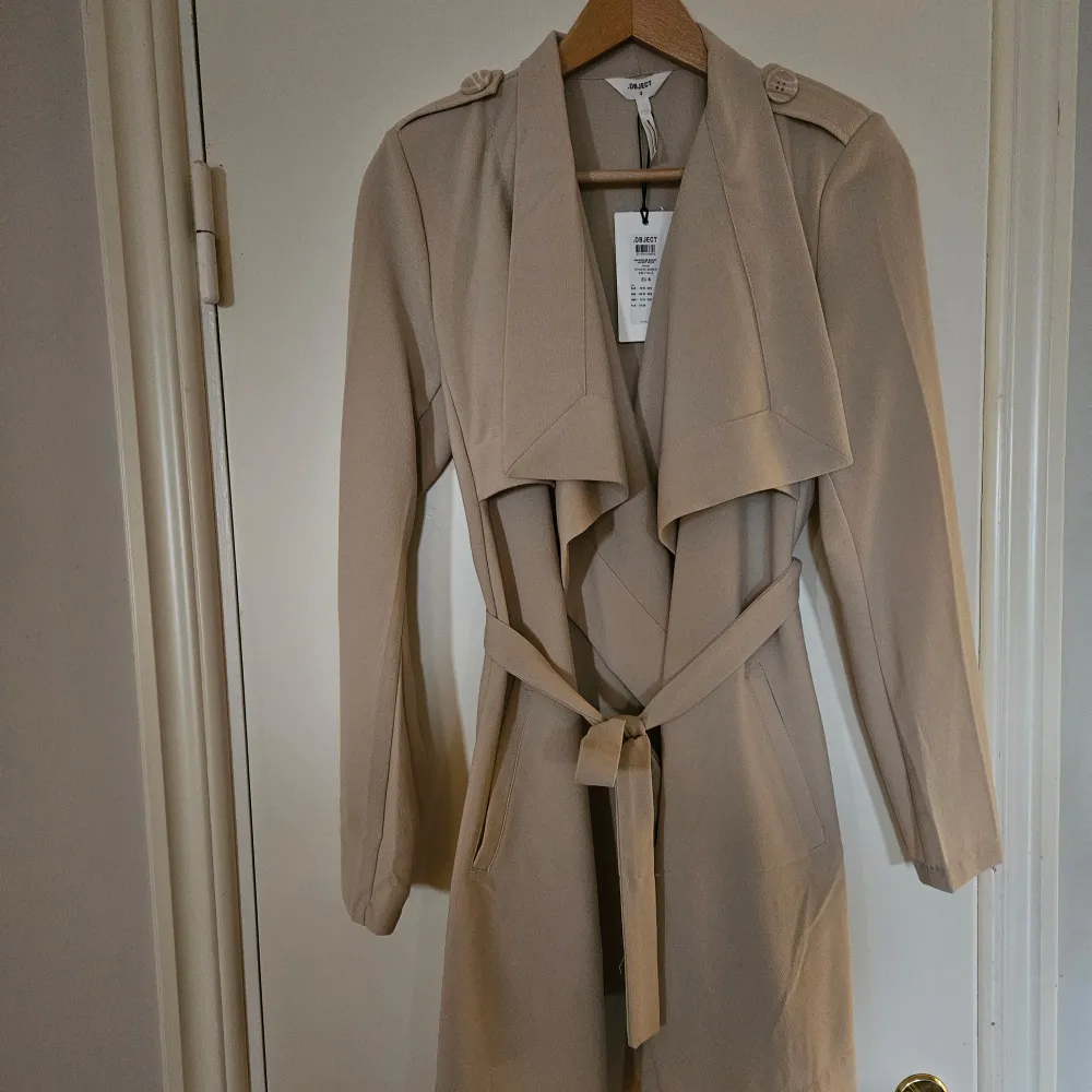  Nytt Säljer en superfin beige trenchcoat i storlek S Den har långa ärmar och ett snyggt knytband i midjan för en elegant look. Jackan har också dekorativa knappar på axlarna och en klassisk krage. Perfekt för höst och vår!. Jackor.