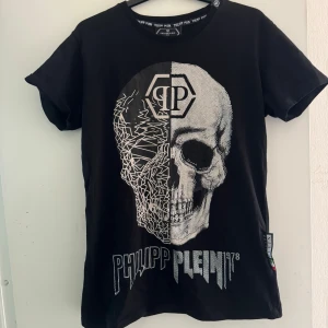 Svart t-shirt med dödskalle från Philipp Plein - Säljer en cool svart t-shirt från Philipp Plein med en stor dödskalle-design på framsidan. T-shirten har korta ärmar och är i nyskick. Perfekt för en edgy look! 💀