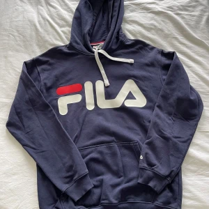 Mörkblå hoodie från FILA - Säljer en snygg och bekväm mörkblå hoodie från FILA. Den har en stor logga på framsidan i vitt och rött. Perfekt för en avslappnad stil och passar bra till både vardag och träning. Den har en klassisk känguruficka och justerbar huva med snören.
