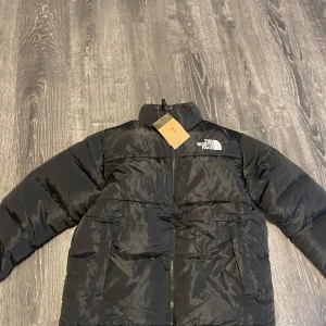 Svart dunjacka från The North Face - Säljer en svart dunjacka från The North Face med dragkedja och hög krage. Jackan har långa ärmar och är perfekt för kalla dagar. Den är stilren och funktionell med märkets logga på bröstet.