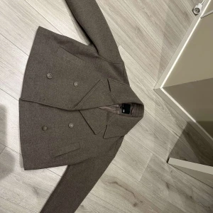 Brun kappa från 157 - Säljer en stilren brun kappa från 157 i storlek S. Jackan har en klassisk dubbelknäppt design med breda slag och långa ärmar. Perfekt för höst och vår!
