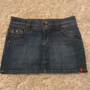 Jeanskjol med fickor - Snygg jeanskjol i klassisk blå denim med dubbla knappar fram och praktiska fickor både fram och bak. Perfekt för en avslappnad stil. Kjolen har hällor för bälte och dekorativa sömmar.