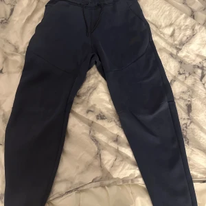 Blå mjukisbyxor från Nike - Snygga blå mjukisbyxor från Nike tech med elastisk midja och snörning. Byxorna har en Nike-logga och praktiska fickor med dragkedja. Perfekta för en avslappnad stil eller träning. Dem är köpta från Zalando för 1100. Pris kan diskuteras och tar byten men föredrar pengar.