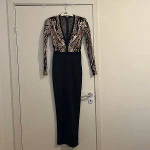 Svart och guld paljettbyxdress i storlek 36 EU från Goddiva - Elegant byxdress från Goddiva i svart med glittrande guldpaljetter. Byxdressen har en djup v-ringning och långa ärmar, perfekt för en glamorös kväll. Den har en figursydd passform som framhäver ens siluett väldigt fint och två fickor framtill. Den är även i nyskick.