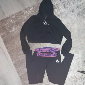 Svart croppad hoodie  + tights från Ellesse - Säljer en svart croppad hoodie från Ellesse med en liten logga på bröstet. Hoodien har långa ärmar och en justerbar huva med dragsko. Perfekt för en avslappnad stil. Byxorna är använda flertal ggr men tröjan endast en gång.