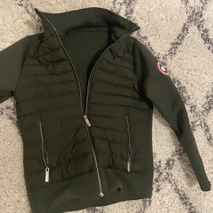 Mörkgrön cardigan från Canada Goose - Säljer en mörkgrön cardigan från Canada Goose med dragkedja och quiltade detaljer. Jackan har långa ärmar och en broderad logotyp på ärmen. Perfekt för kyligare dagar. Storlek: S.