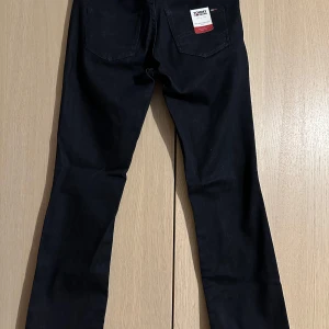 Svarta jeans från Tommy Jeans - Snygga svarta jeans från Tommy Jeans med en klassisk straight passform. De är midwaist och har en stilren design med fem fickor och en läderpatch med logotyp på baksidan. Perfekta för en avslappnad look.
