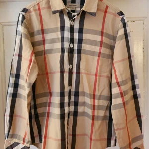 Burberry skjorta - Burberryskjorta i klassisk, rutig design i beige, svart, vitt och rött. Mycket bra skick överlag, dock är den lite sliten längst ner på manchetterna (se bild!), ej så synligt när man bär den. Tvättrådslappen är helt urtvättad, men extraknappen sitter kvar. Storlek 10 år, 138 cm. 