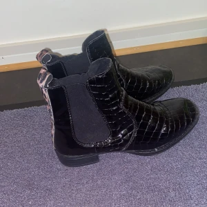 Svarta boots från Rieker - Snygga svarta boots från Rieker med krokodilpräglat mönster och leoparddetaljer på hälen. De har en elastisk sida för enkel påtagning och en låg klack för bekväm användning. Perfekta för att ge en stilfull touch till din outfit.