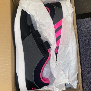 Svarta och rosa sneakers från Adidas - Snygga svarta sneakers med rosa detaljer från Adidas. Skorna har kardborreband för enkel på- och avtagning. De har en vit sula och tre rosa ränder på sidan. ALDRIG ANVÄNDADE