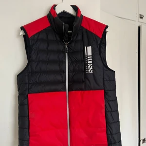 Svart och röd dunväst från Hugo Boss - Snygg svart och röd dunväst från Hugo Boss med dragkedja framtill. Västen har en hög krage och är vattenavvisande samt lättviktig. Perfekt för kyligare dagar.
