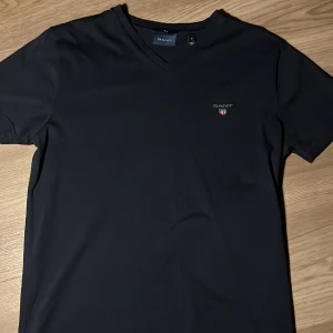 Svart V-cut  t-shirt från GANT - Snygg svart t-shirt från GANT med v-ringning och diskret logga på bröstet. Perfekt för en stilren look.