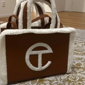 UGG x Telfar brun väska - Säljer en väldigt rymlig snygg brun UGG x Telfar väska med vit pälsdetalj. Väskan har dubbla korta och långa handtag/axelremsband och ett stort Telfar-logotyp på framsidan. Finns även ett extra fack med dragkedja på insidan. Helt oanvänd🌟
