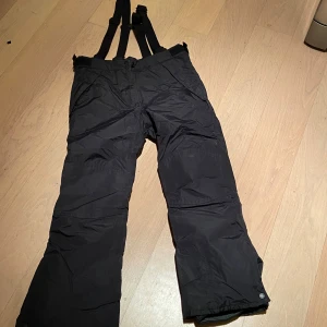 Svarta täckbyxor från Everest stl 146 nyskick  - Svarta täckbyxor från Everest med justerbara hängslen och elastiska benslut med snölås. Byxorna stängs med knapp och dragkedja. 