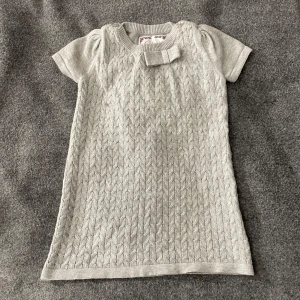 Grå stickad barn klänning från Skill - Söt grå stickad klänning från Skill med korta ärmar och ett fint flätmönster. Klänningen har en dekorativ rosett vid halsen och är perfekt för kyligare dagar. Storlek 80.