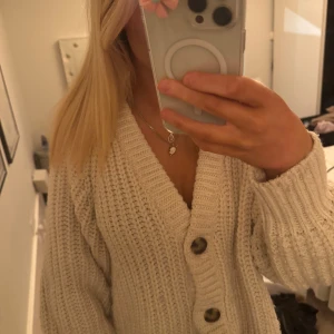 Beige stickad kofta - Mysig beige stickad kofta med stora knappar framtill. Perfekt för kyliga dagar och ger en avslappnad look. Koftan har en v-ringad design och långa ärmar.