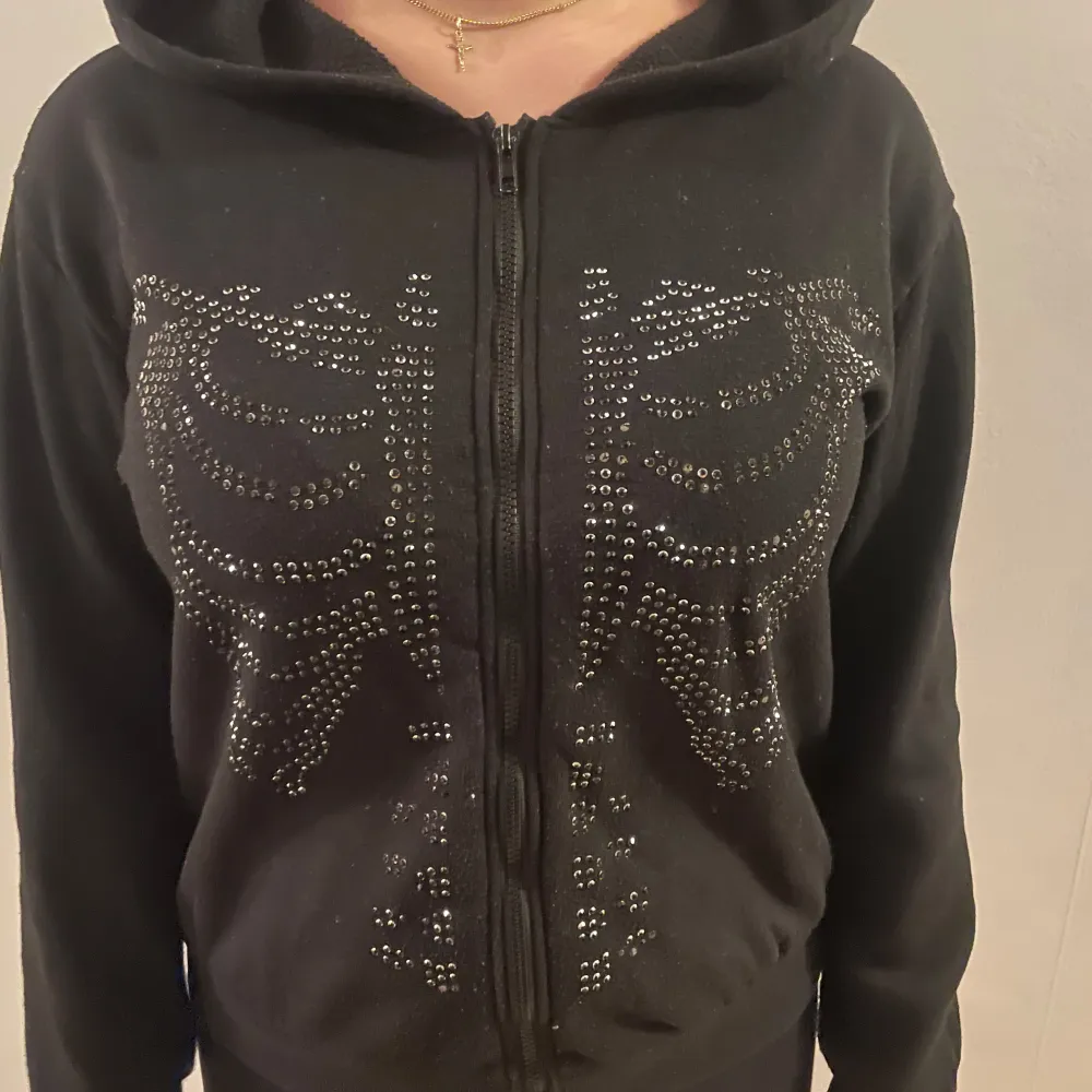 Snygg svart hoodie med dragkedja och paljettdetaljer i form av ett skelettmönster på framsidan. Perfekt för en cool och avslappnad stil. Den har långa ärmar och en bekväm passform.. Neuletakit & Villapaidat.