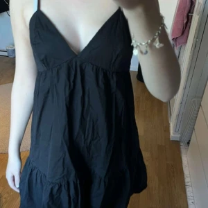 Svart klänning med öppen rygg - Snygg svart klänning med v-ringning och öppen rygg. Den har smala axelband och en volangdetalj vid fållen. Perfekt för en somrig look eller en utekväll. Klänningen har en avslappnad passform och knytning i ryggen för extra stil. Använd endast en gång!