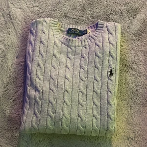 Grå stickad tröja från Ralph Lauren - Säljer en stilren grå stickad tröja från Ralph Lauren med klassiskt kabelstickat mönster. Tröjan har en broderad logotyp på bröstet och långa ärmar. Perfekt för en avslappnad men ändå elegant look. Passar till allt