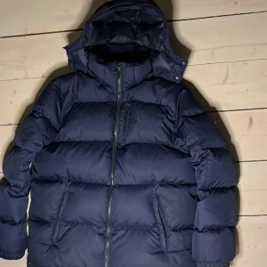 Mörkblå dunjacka från Ralph Lauren - Säljer en snygg mörkblå puffer jacket från Ralph Lauren. Jackan har en dragkedja framtill och en broderad logotyp på bröstet. Perfekt för kalla dagar med sin vadderade design och huva.