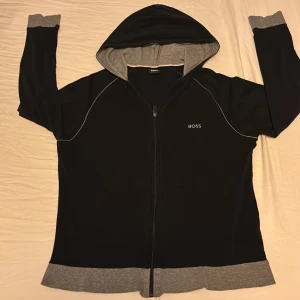 Svart hoodie från Boss - Säljer en svart hoodie frånHugo  Boss med gråa detaljer. Den har en dragkedja framtill och en skön och snygg luva. Storlek M och ett väldigt bra skick men varan är andvänd vid ett få antal gånger. Varan kostar Ca 900 i butik. Så ett väldigt bra pris.