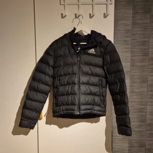 Svart dunjacka från Adidas - Säljer en svart dunjacka från Adidas med dragkedja och långärmad design. Jackan har en klassisk pufferstil och är perfekt för kyligare väder. Adidas-loggan syns tydligt på bröstet. Perfekt för dig som vill ha en stilren och funktionell jacka.