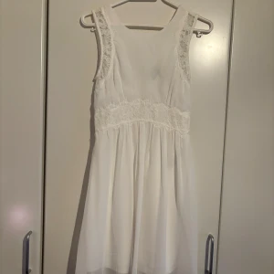 Vit klänning från Vero Moda - Säljer en elegant vit klänning från Vero Moda med spetsdetaljer på axlarna och midjan. Klänningen har en v-ringad design och är ärmlös, vilket ger en luftig och stilren look. Perfekt för sommarens alla tillfällen! 🌸 