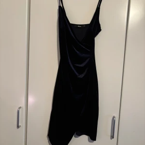 Svart klänning från Bershka - Elegant svart klänning från Bershka med smala axelband och omlott-design. Perfekt för en stilren look med en touch av glamour. Klänningen har en v-ringad hals och är tillverkad i ett glansigt material som ger en lyxig känsla.