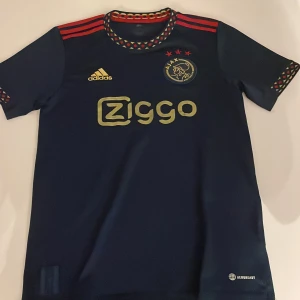 Mörkblå Ajax fotbollströja från Adidas - Snygg mörkblå Ajax fotbollströja med guldiga detaljer och röd dekor. Tröjan har korta ärmar och ett mönster runt halsen och ärmsluten. Perfekt för alla Ajax-fans!