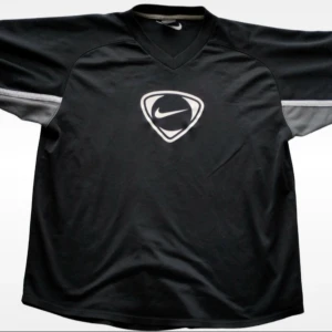 Svart sporttröja från Nike 90’s Vintage  - Säljer en svart sporttröja från Nike med ett stort vitt logotryck på bröstet. Tröjan har korta ärmar och en v-ringad hals. Perfekt för sportiga aktiviteter eller en avslappnad stil. 90's Vintage. Mått: armhåla-armhåla 60 cm. Längd 70cm armlängd 26 cm axel-armhåla 30 cm