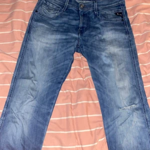 Blå jeansbyxor från Replay  - Snygga blå jeansbyxor från replay med en klassisk femficksdesign och knappgylf. De har en lätt slitning på ena benet som ger en cool, avslappnad look. Perfekta för en casual stil.