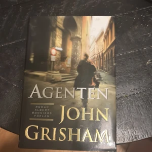 Agenten - Säljer boken 'Agenten' av John Grisham, utgiven av Albert Bonniers Förlag. Omslaget visar en mystisk figur i en stadsmiljö, vilket ger en spännande och dramatisk känsla. Perfekt för dig som gillar thrillers och spionhistorier.