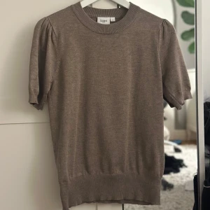 Beige/brun tröja från Saint - Säljer en stilren beige tröja från Saint med korta ärmar och rund halsringning. Tröjan har en ribbad kant vid ärmslut och nederkant, vilket ger en snygg passform. Perfekt för skola eller finare evenemang.