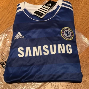 Blå Chelsea fotbollströja från Adidas - Vi säljer nu ett helt nytt (oanvänt) Chelsea fotbollskit (tshirt och shorts ). Passar 10-11 år. Detta är en julklapp som inte passade mottagaren som håller på annat lag.. Men icke desto mindre perfekt för ett fan av Chelsea FC! 💙