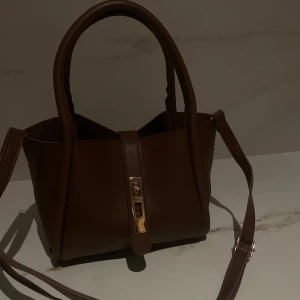 Brun handväska med gulddetaljer - Snygg brun handväska i skinn imitation med gulddetaljer. Väskan har en elegant design med ett praktiskt handtag och en justerbar axelrem. Väskan är oanvänd 