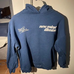 Blå hoodie med tryck - Säljer en blå hoodie med tryck på både fram- och baksidan. Texten 'new mind illusion' och 'UNREAL 2'.  Hoodien har långa ärmar och en klassisk huva.