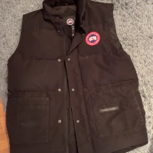 Svart dunväst från Canada Goose - Säljer en svart dunväst från Canada Goose med märkets logga på bröstet. Västen har dragkedja och knappar framtill samt två stora fickor. Perfekt för kyliga dagar.
