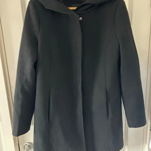 Svart kappa med huva - Snygg svart kappa med huva, perfekt för kyliga dagar. Den har en stilren design med långa ärmar och knäppning framtill. Passar bra till både vardag och mer uppklädda tillfällen.