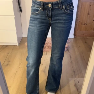 Blå jeans från Gina Tricot - Snygga blå jeans från Gina Tricot med bootcut-stil och klassisk femficksdesign. De har en knapp och dragkedja framtill samt dekorativa fickor med knappar bak. Perfekta för en avslappnad look.