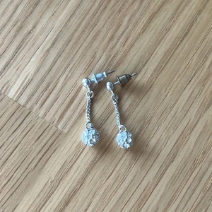 Silverfärgade örhängen med glittrande detaljer - Silverfärgade örhängen med hängande kedja och strassklädd boll. Väldigt lätta och därför bekväma och enkla att bära. Säljer då jag inte använder silver. Tyvärr finns inte orginalplupparna så därför är de missmatchande men detta påverkar inte användningen. Örhängena är noga tvättade och disinfekterade. Märket är okänt.
