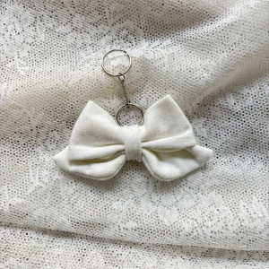 Vit rosett i tyg - Charmig vit rosett i tyg. Perfekt som en stilren accessoar för att hålla dina nycklar samlade. Rosetten är gjord av ett mjukt material och fäst vid en silvrig metallkedja med en ring.