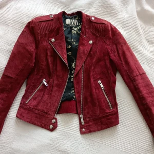 Röd mockajacka från Jofama by Marie Sernholt  - Snygg vinröd biker-jacka i mockafrån Jofama med dragkedja och knappar. Jackan har långa ärmar och ett stilrent blommigt foder. Perfekt för att ge din outfit en cool touch Säljes pga det egentligen är fel storlek för mig.