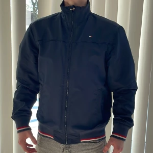 Mörkblå jacka från Tommy Hilfiger - Snygg mörkblå jacka från Tommy Hilfiger med dragkedja och ribbade muddar i rött, vitt och blått. Jackan har en hög krage och långa ärmar, perfekt för en stilren look, Nypris 1899kr. Klassisk design med subtil logga på bröstet. Bra pris just nu på en sån här stilren jacka 🧥 